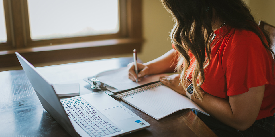 woman sitting at desk writing in a notebook with open laptop